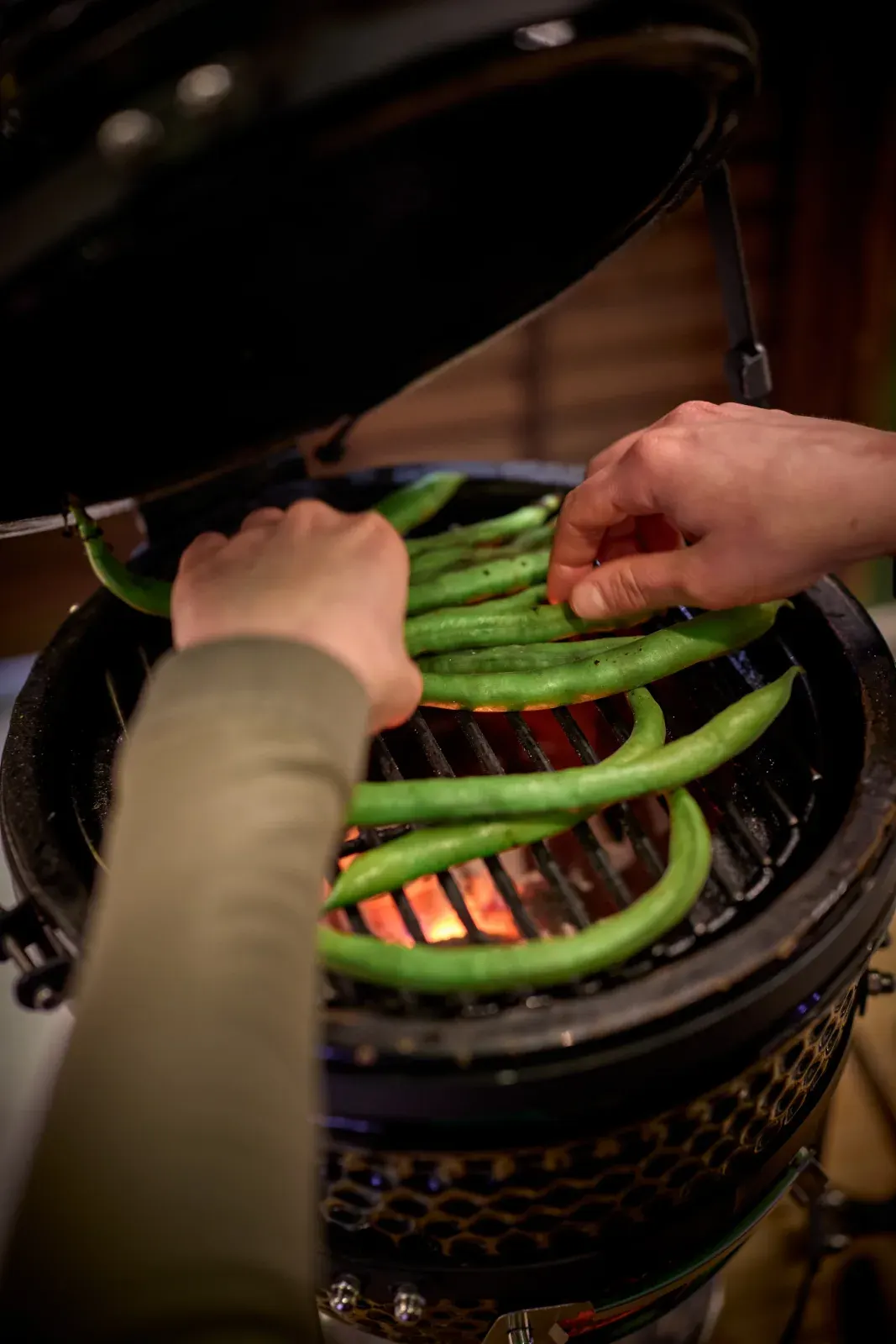 Las manos del equipo colocan vainas verdes sobre la parrilla, con la brasa encendida debajo