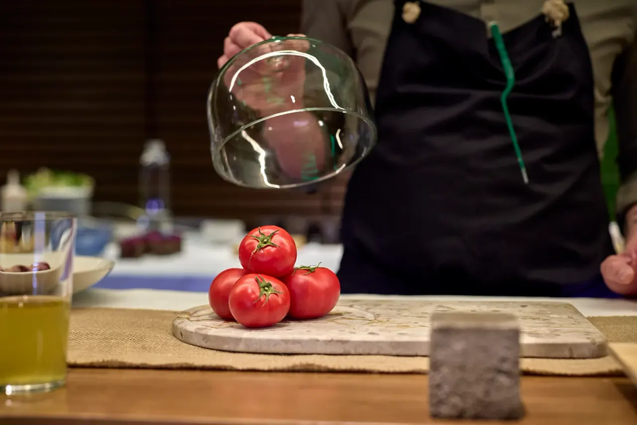 Tres tomates bajo una campana de cristal que una mano levanta, sobre un disco de mármol